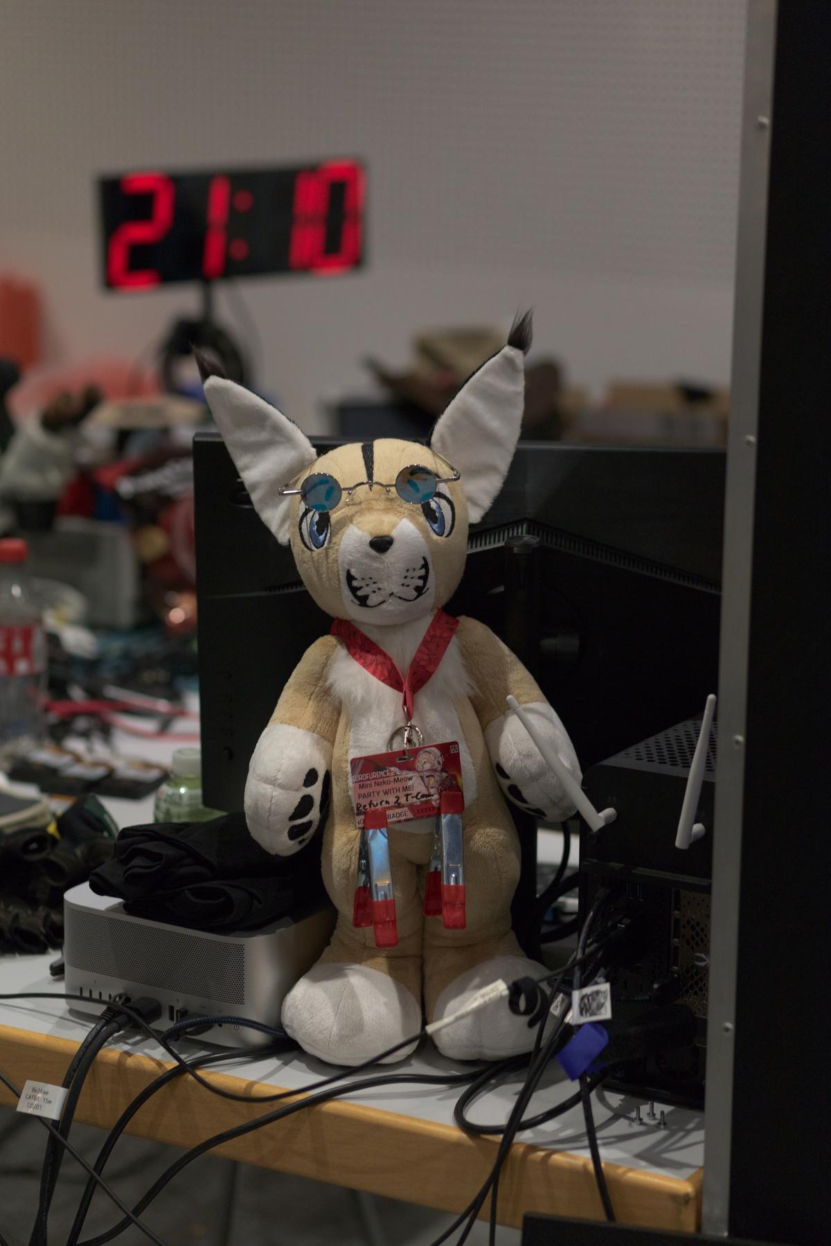 A caracal plush toy stands on a table in front of a monitor. It wears silver glasses with blue lenses and an EF29 non-Badge: Mini Neok-Meow PARTY WITH ME! Return 2 T-Com. There's a large 7 segment display in the background showing 21:10.