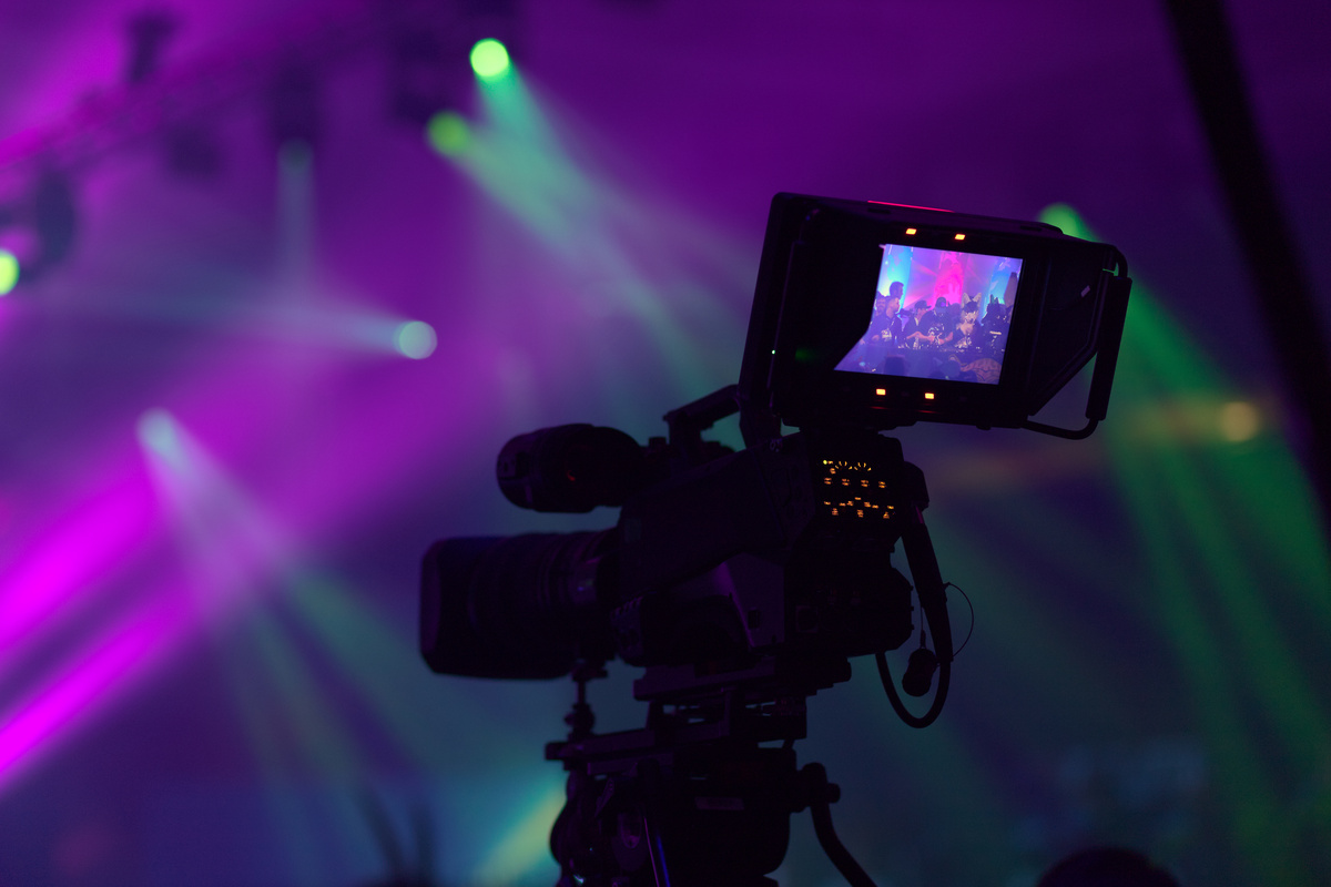 Picture of a camera in Hall H. The cameras monitor is in focus and shows part of the stage with dancing furries and there are some out of focus lights in the background.