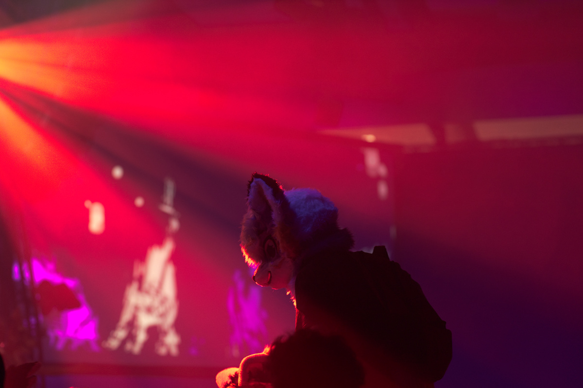 Yet another fursuiter is sitting on the shoulders of another person and is illuminated by some red light.