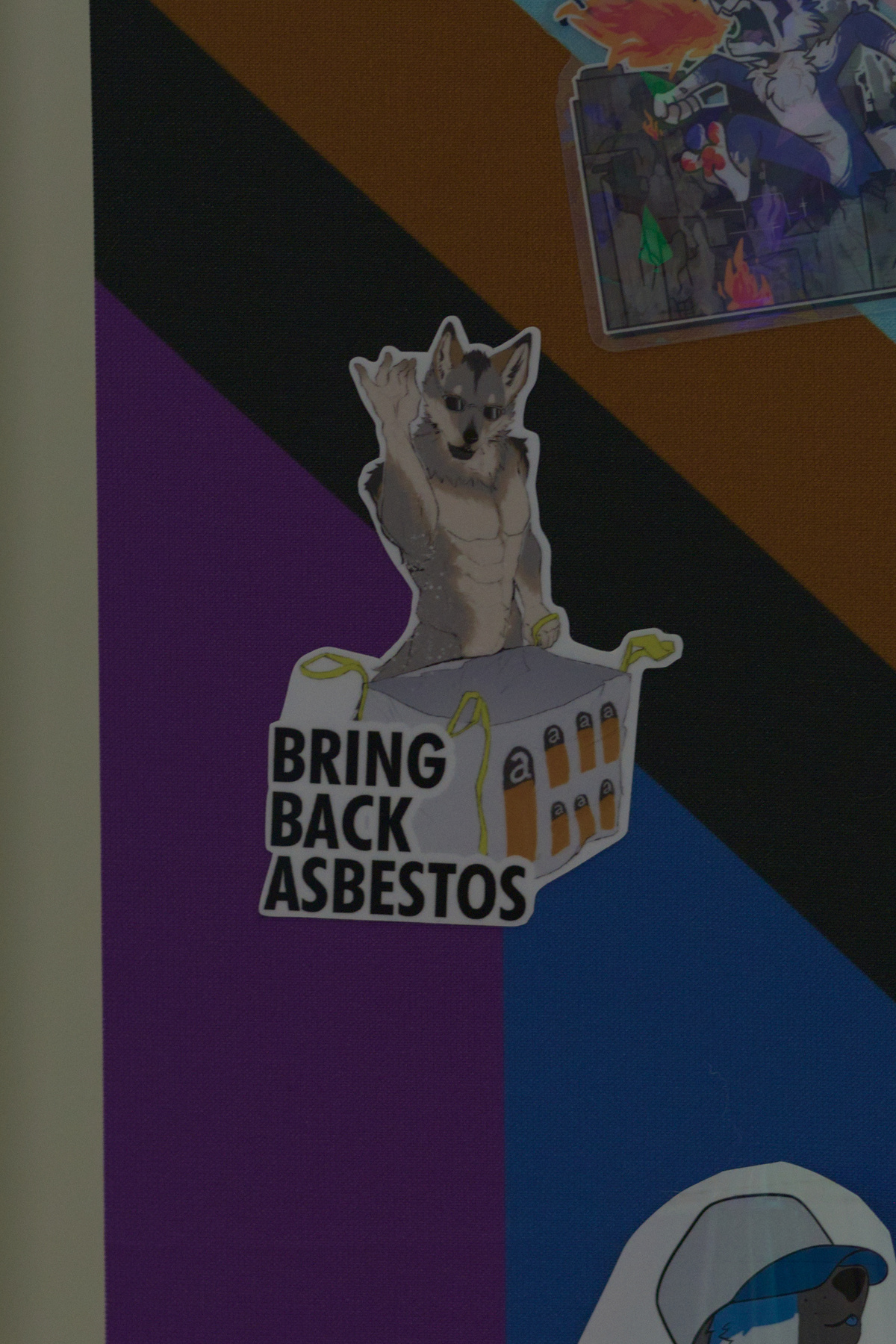 Sticker wall: a 'bring back asbestos' sticker depicting a wolf with sunglasses drizzling a pinch of asbestos into a labeled bag